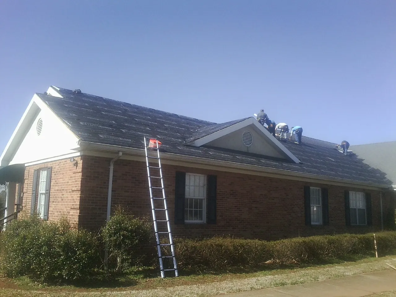 Crew tearing shingles off a brick ranch roof with a ladder set — Emergency Roof Repair work by our crew in Enid