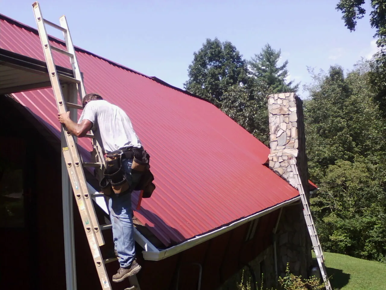Roofer on a ladder beside a red metal roof and stone chimney — Soffit & Fascia Repair by Roofing Brothers in Enid